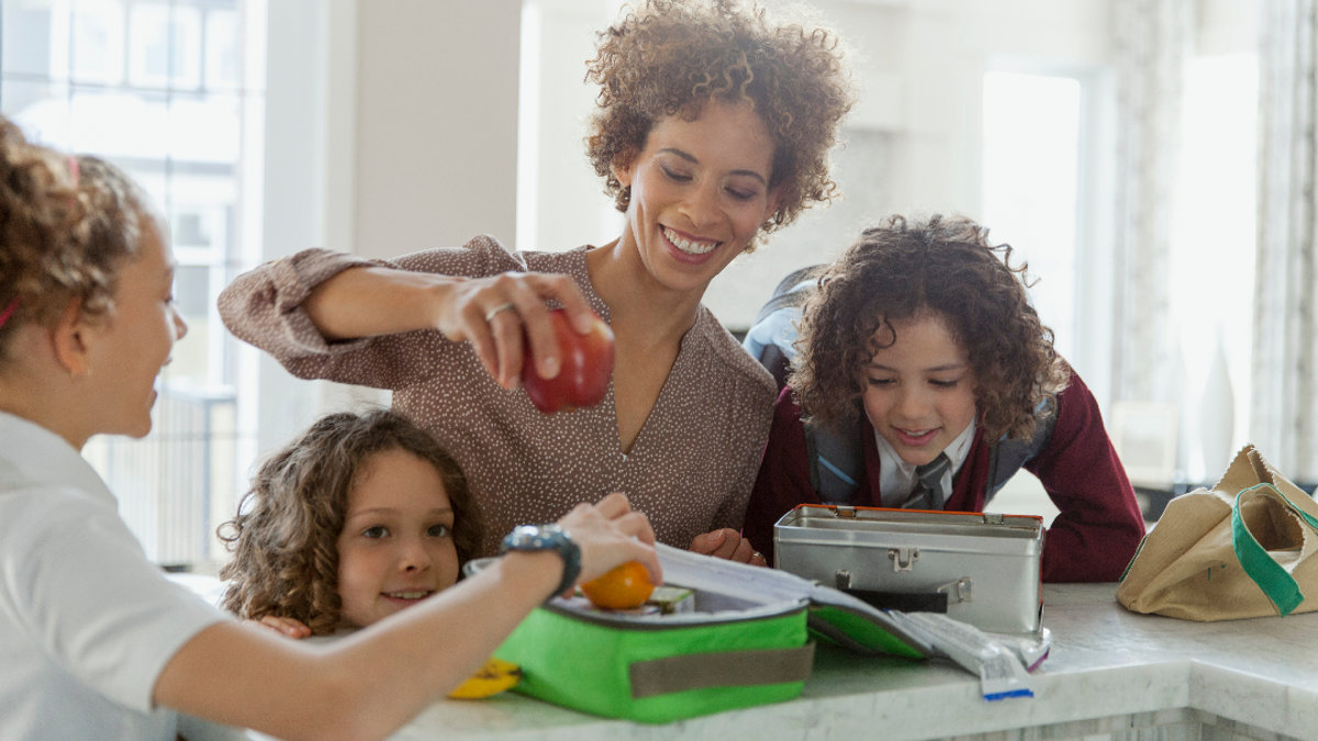 Adult woman packing a child's lunch box with her 3 children surrounding her at the kitchen worktop smiling
