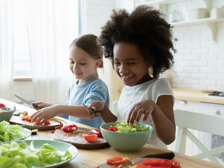 Two young girls sitting at kitchen table cutting and cutting up salad.