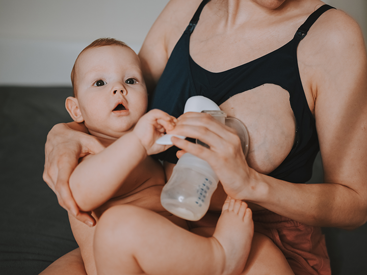 Woman using a pump in one hand to express milk whilst holding her baby in her other arm