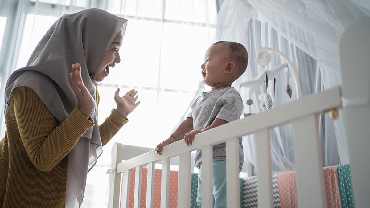 Woman holding her hands wide open and smiling at a young boy standing up in a cot