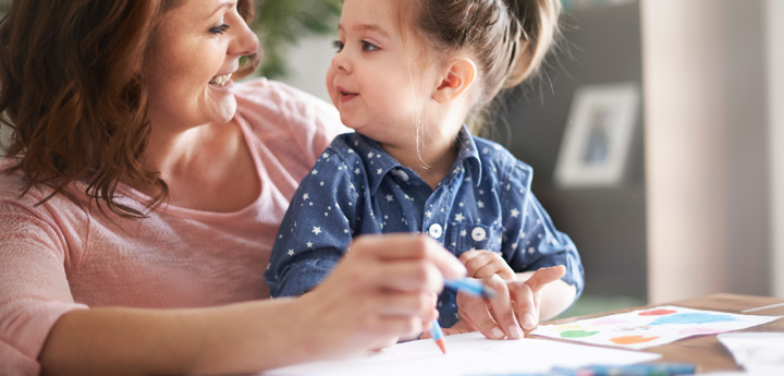 Young girl sitting on an older woman's lap, smiling at each other. They are both holding a crayon and a piece of paper is in front of them on a table