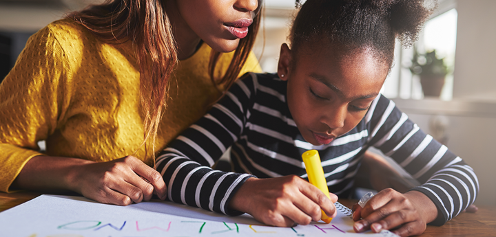child writing ABCs on large paper at a table with adult woman sitting next to her peering over her shoulder