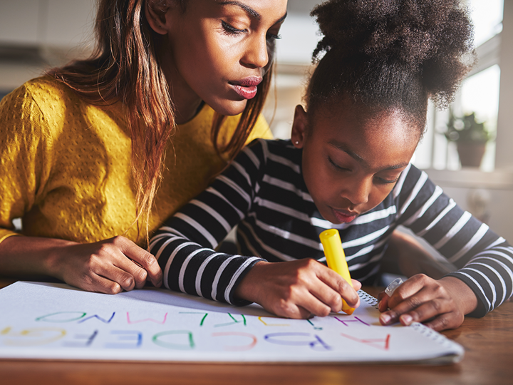 child writing ABCs on large paper at a table with adult woman sitting next to her peering over her shoulder