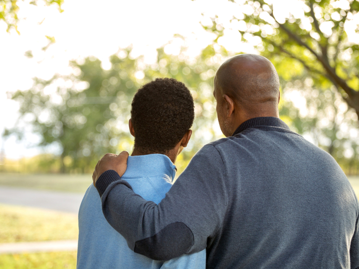 Teenage boy and dad standing with their back to the camera. The dad has his arm around his son.