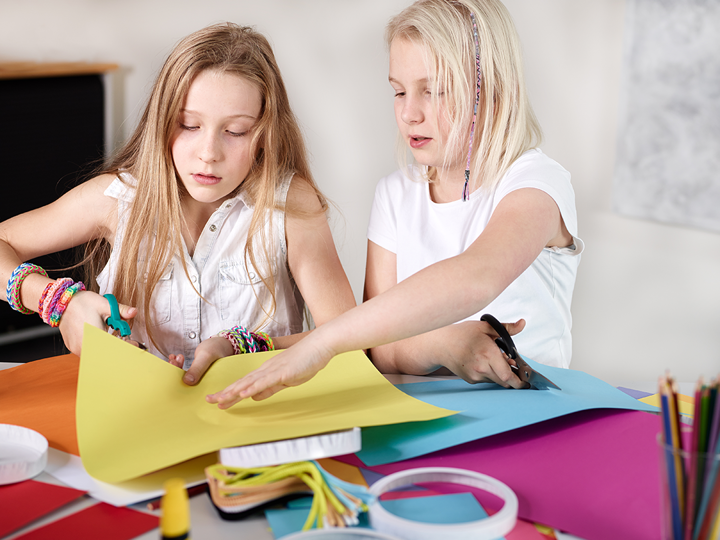Two girls sitting at a table both cutting coloured paper with scissors