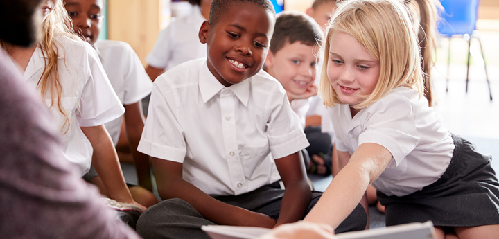 Children In A Class Looking At A Book Held By A Teacher