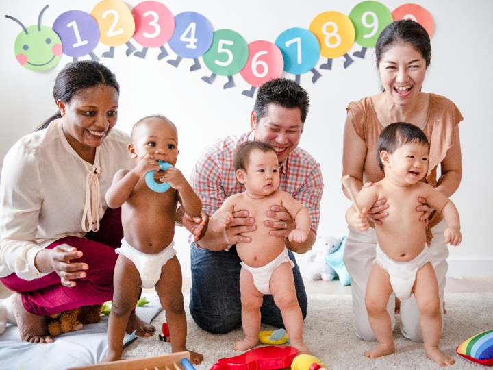 3 babies standing in a row in nappies, each being supported by an adult kneeling behind them
