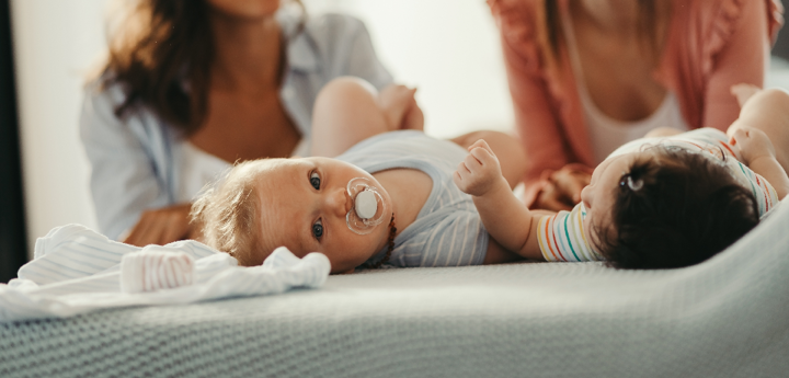 Two women kneeling behind their babies, which are both lying on a bed on their back
