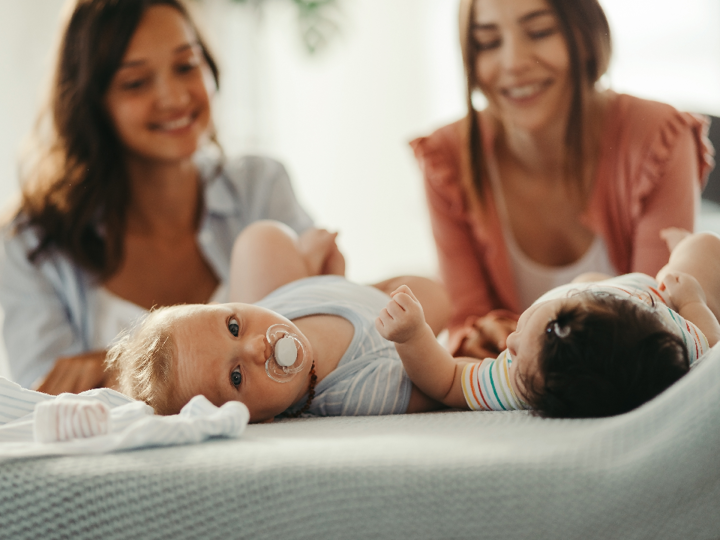 Two women kneeling behind their babies, which are both lying on a bed on their back