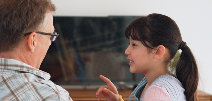 A young girl sitting on the sofa with an adult man, the girl is talking to him with one finger lifted as she speaks