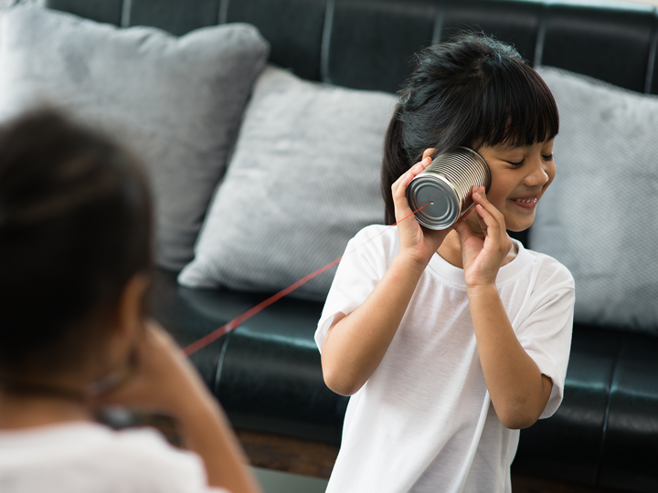 Two girls playing telephone with tin cans and string. Background is a grey sofa. Girl in foreground listening to tin can and smiling