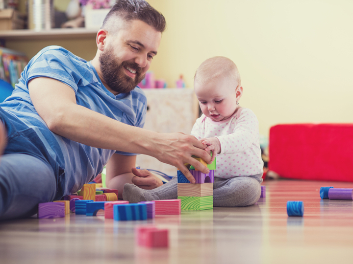 Baby sitting on the floor playing with blocks in front of them. Dad smiling and lying next to them on the floor also playing with the blocks.