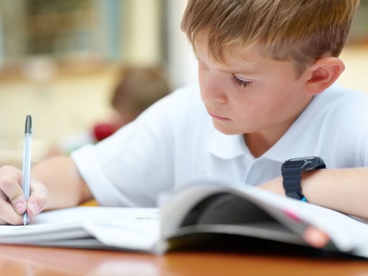 Close up photograph of young blond haired boy doing school work at a desk in a classroom. 