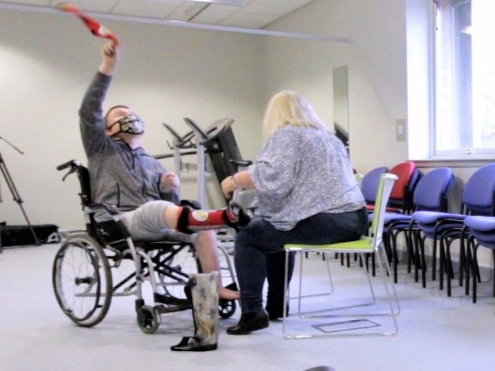 Young person in a physio clinic sitting in a wheelchair whilst an adult sits opposite them. The young person's foot is resting on the adult's knee.