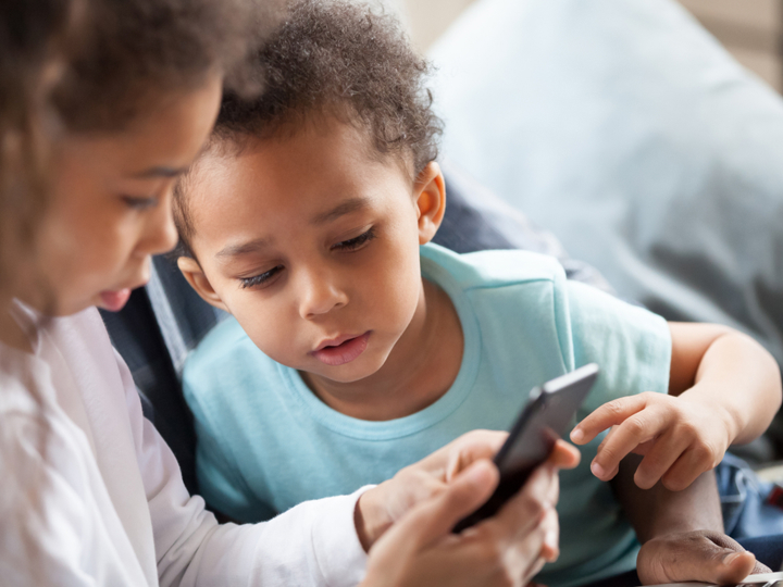 Young girl on mobile phone with a younger boy peering over her shoulder to look at the mobile phone.