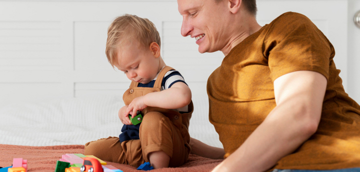 Father And Son Sitting On A Bed Playing With Coloured Blocks And Toy Cars
