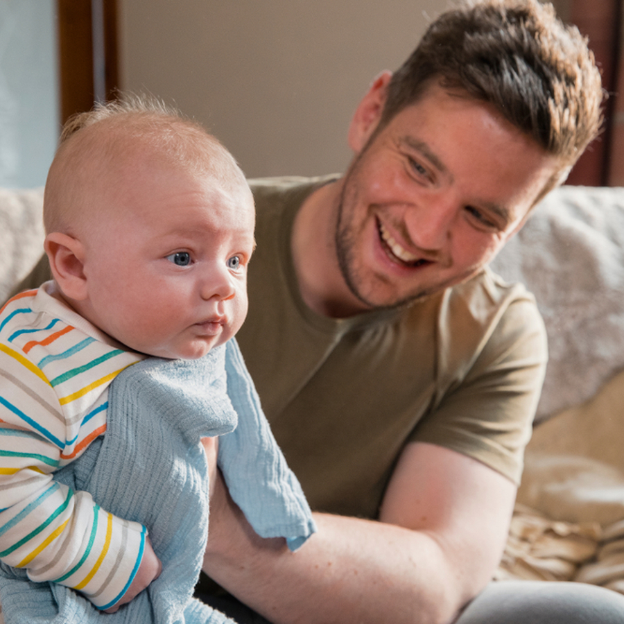 Dad sitting on the edge of sofa holding a baby and patting them on the back