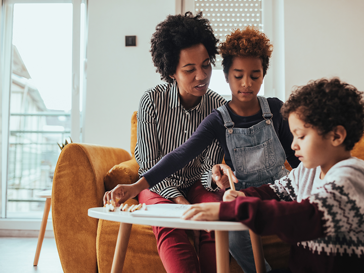 Adult woman and child sitting on a sofa looking at a young boy sitting on the floor writing on a piece of paper on a coffee table in front of them