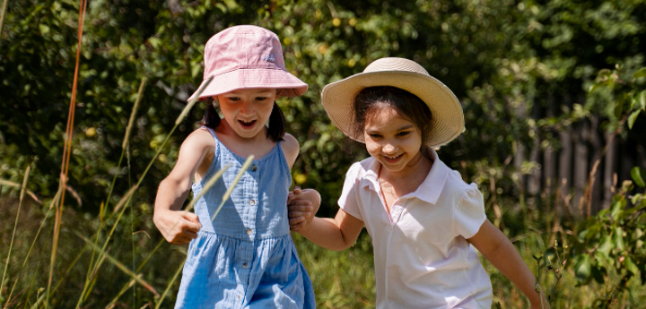 Two young girls running through grass in the sun wearing sun hats