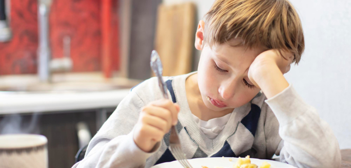Young boy sitting at the kitchen table looking miserable. Holding a fork in one hand and his head is resting in his palm of the other hand.