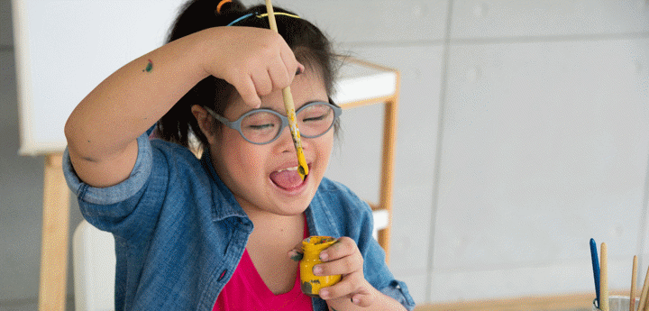 Young girl with glasses painting, sitting at a table dipping a paintbrush into a paint pot and smiling.