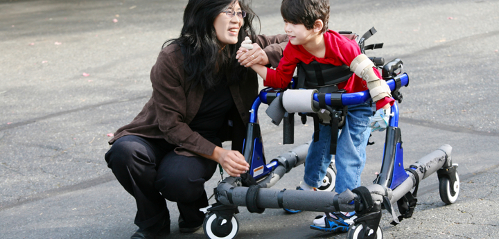 Adult woman crouching next to young boy with walking support. Woman is holding the boys hand.