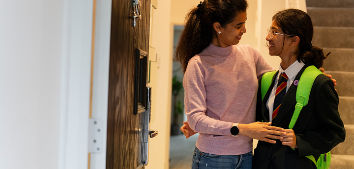 Adult woman smiling and hugging smiling teenage girl in school uniform with lime green backpack. Inside a home with door open.