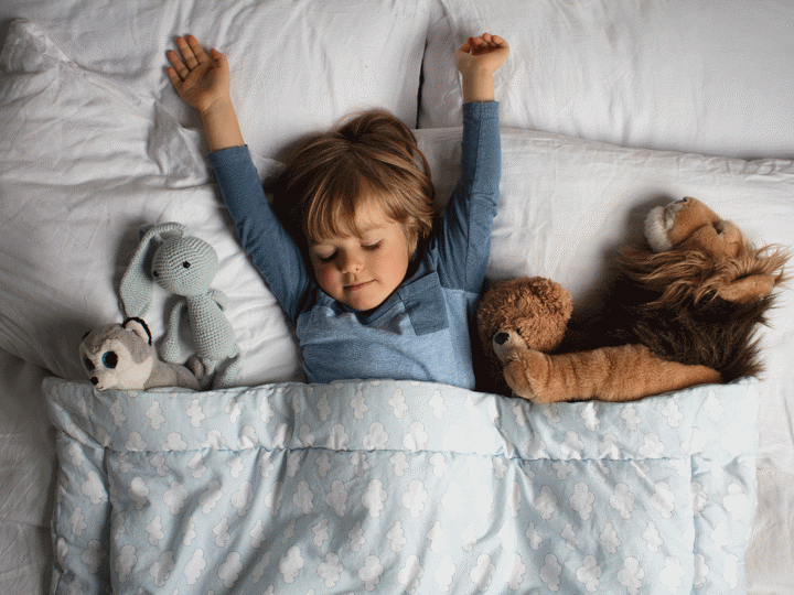child lying in bed with their arms outstretched above them on the pillow and stuffed animals under the duvet either side of them